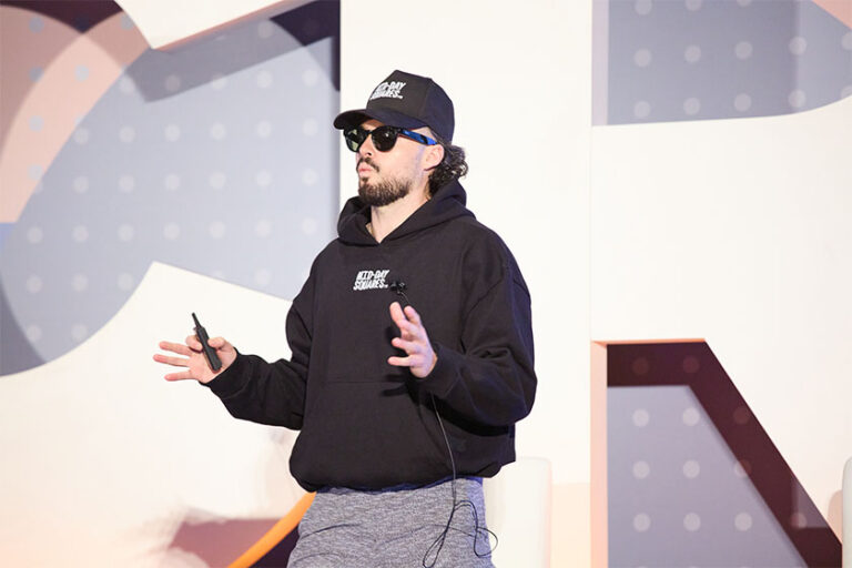 A man in a black sweatshirt and hat speaking on stage